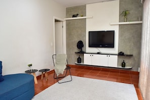 Ein modernes Wohnzimmer mit einem Flachbildfernseher an einer grauen Wand, einem blauen Sofa, einem kleinen Tisch mit einer Pflanze und einem Sessel mit Überwurfdecke.
