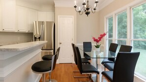 Une cuisine moderne avec des meubles blancs, un réfrigérateur en acier inoxydable, une table à manger en verre avec des chaises noires et une fenêtre donnant sur les arbres.