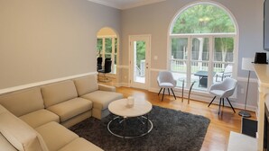 Un salon spacieux avec un canapé d’angle beige, une table basse ronde et un ventilateur de plafond. On y trouve deux chaises blanches et une petite table près d’une grande fenêtre donnant sur un paysage verdoyant.