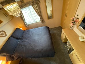 A bedroom with a bed, a dresser, a mirror, a window with curtains, and a chandelier. - Walsh's Holiday Park - The Sapphire (Skegness)