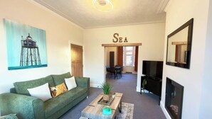 Ein Wohnzimmer mit einem grünen Sofa, einem Couchtisch, einem Fernseher und einem Gemälde eines Leuchtturms.