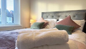 A neatly made bed with a tufted headboard, a bedside lamp, and a window with blinds. - Driftwood Bay - Coastal Escape (Lancashire)