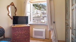 A room with a television on a dresser, a window with curtains, a radiator, and a view of a hillside town. - Palazzo Nobili (Capri)