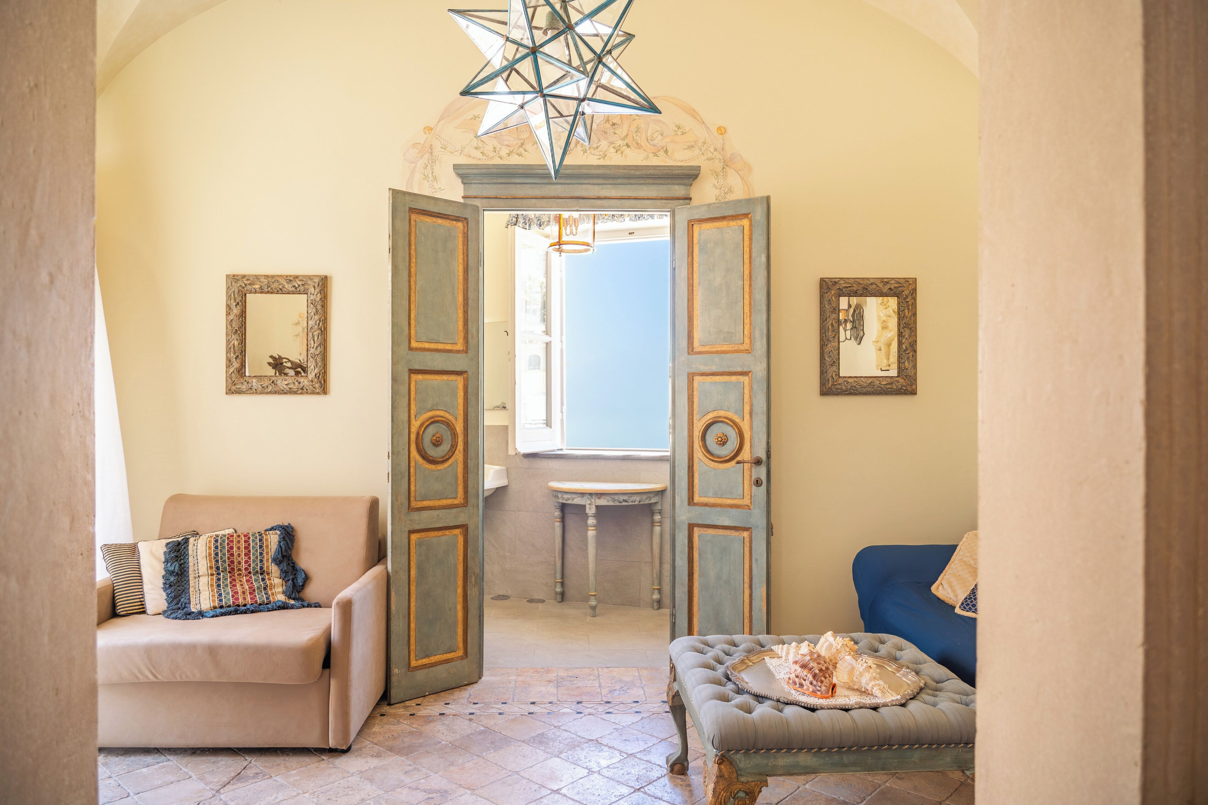 An open doorway leading to a bathroom with a window, a beige sofa, a blue armchair, and a decorative star-shaped light fixture.