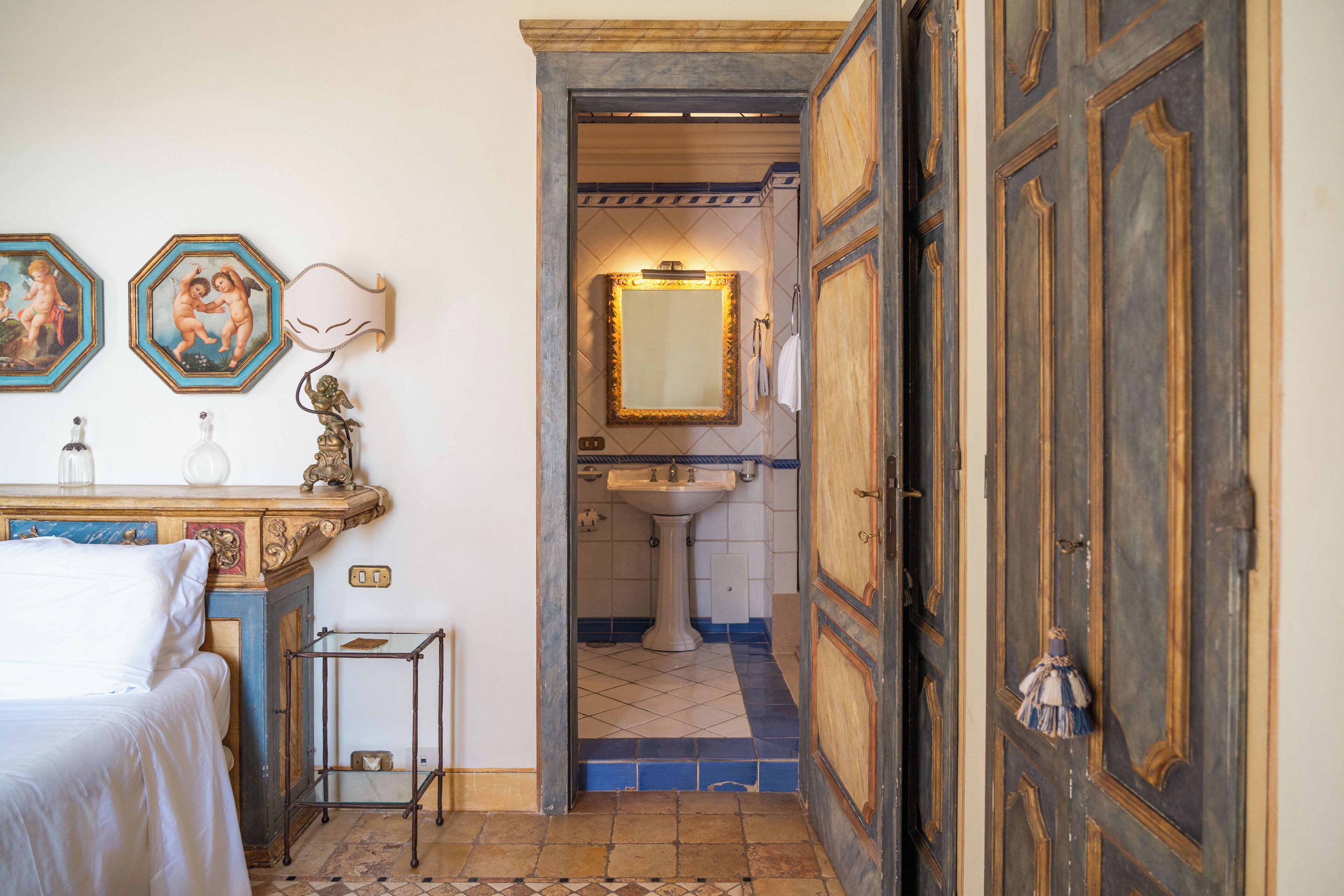 Una habitación con una cama, una mesita, un espejo y una puerta que da a un baño con lavabo y suelo de baldosas.