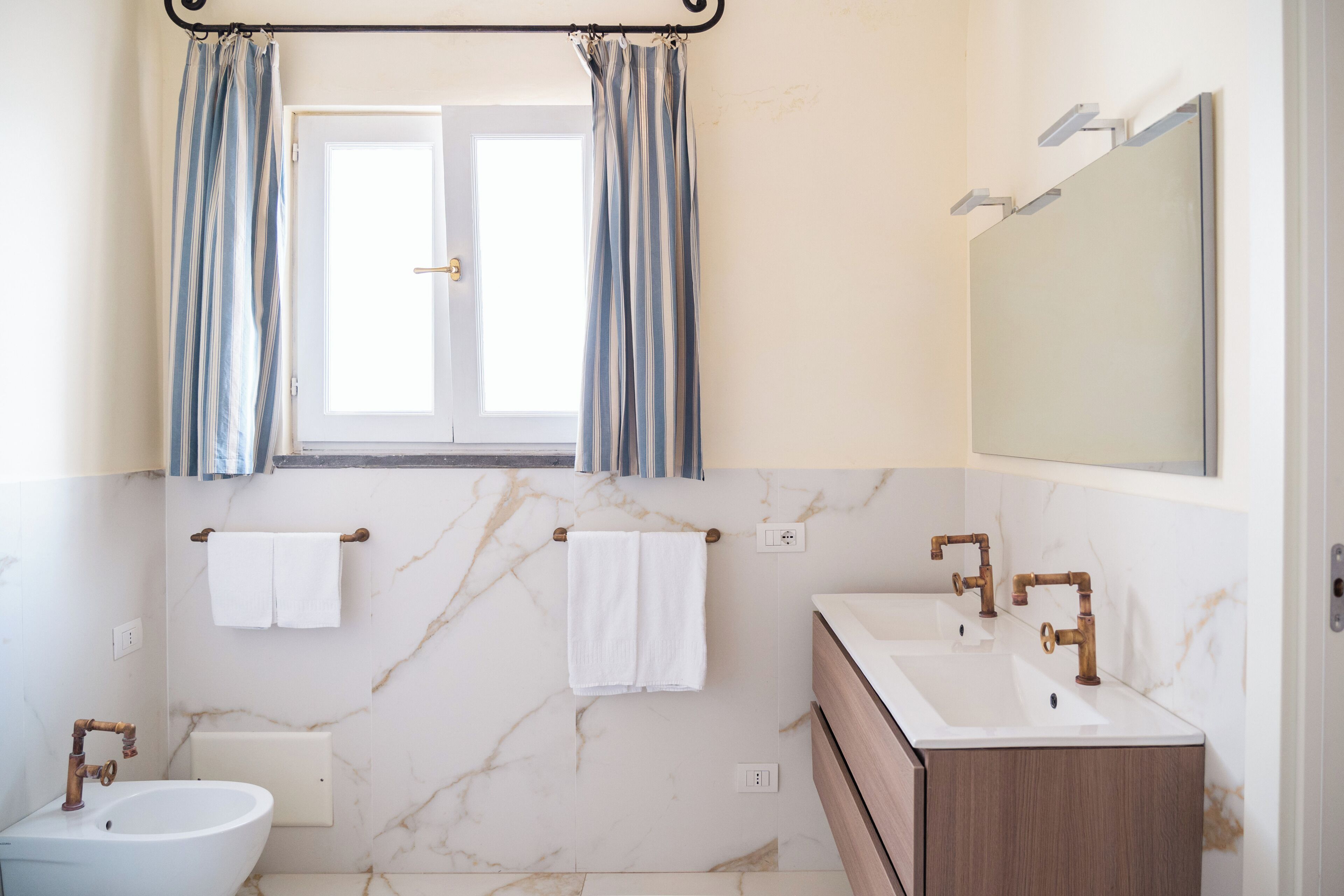 A bathroom with a window, two sinks, a bidet, marble walls, and a mirror.