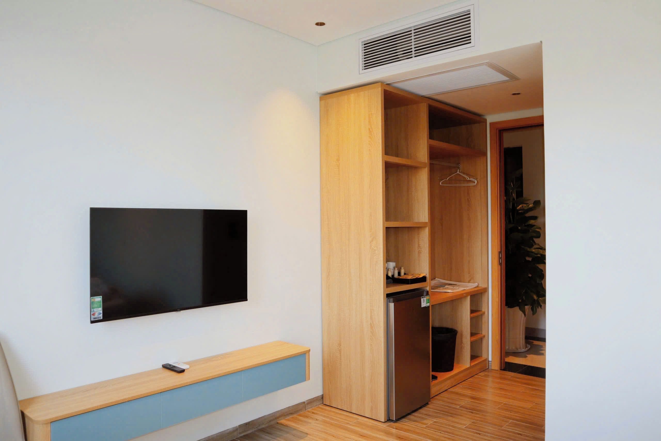 Una habitación moderna con un televisor de pantalla plana montado en la pared, un armario de madera con las puertas abiertas y un pequeño banco con un control remoto.