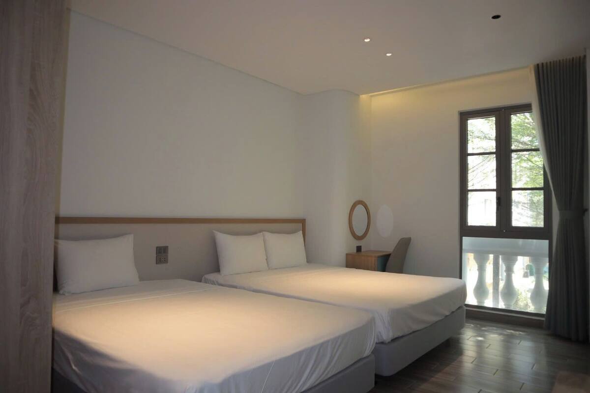 Habitación de hotel con dos camas, cabecera de madera, mesita de noche pequeña con espejo y ventana con cortinas.