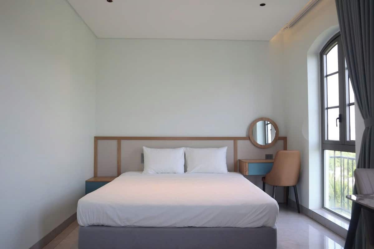 Una habitación de hotel con una cama grande, un espejo redondo, una mesita de noche, una silla y una ventana con cortinas.