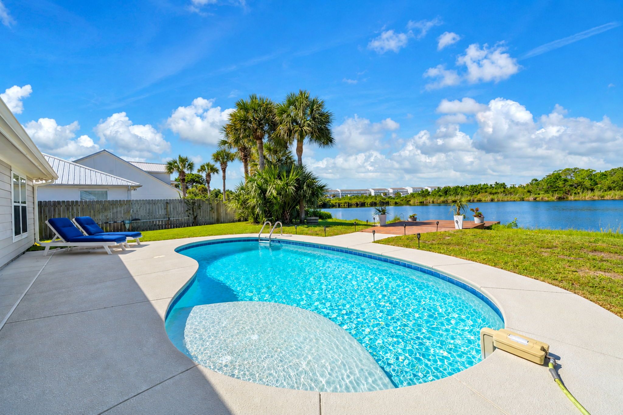 A swimming pool with a wooden deck, lounge chairs, and a view of a lake and buildings in the background.