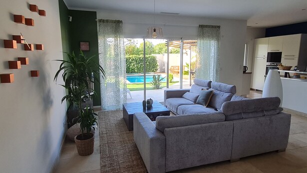 Sala de estar moderna com sofá cinzento, mesa de centro e uma grande planta em vaso. Há uma porta de vidro deslizante que dá para uma área de piscina.