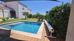 Una piscina rectangular con una cubierta de madera, rodeada por un seto y un edificio blanco con techo de tejas.