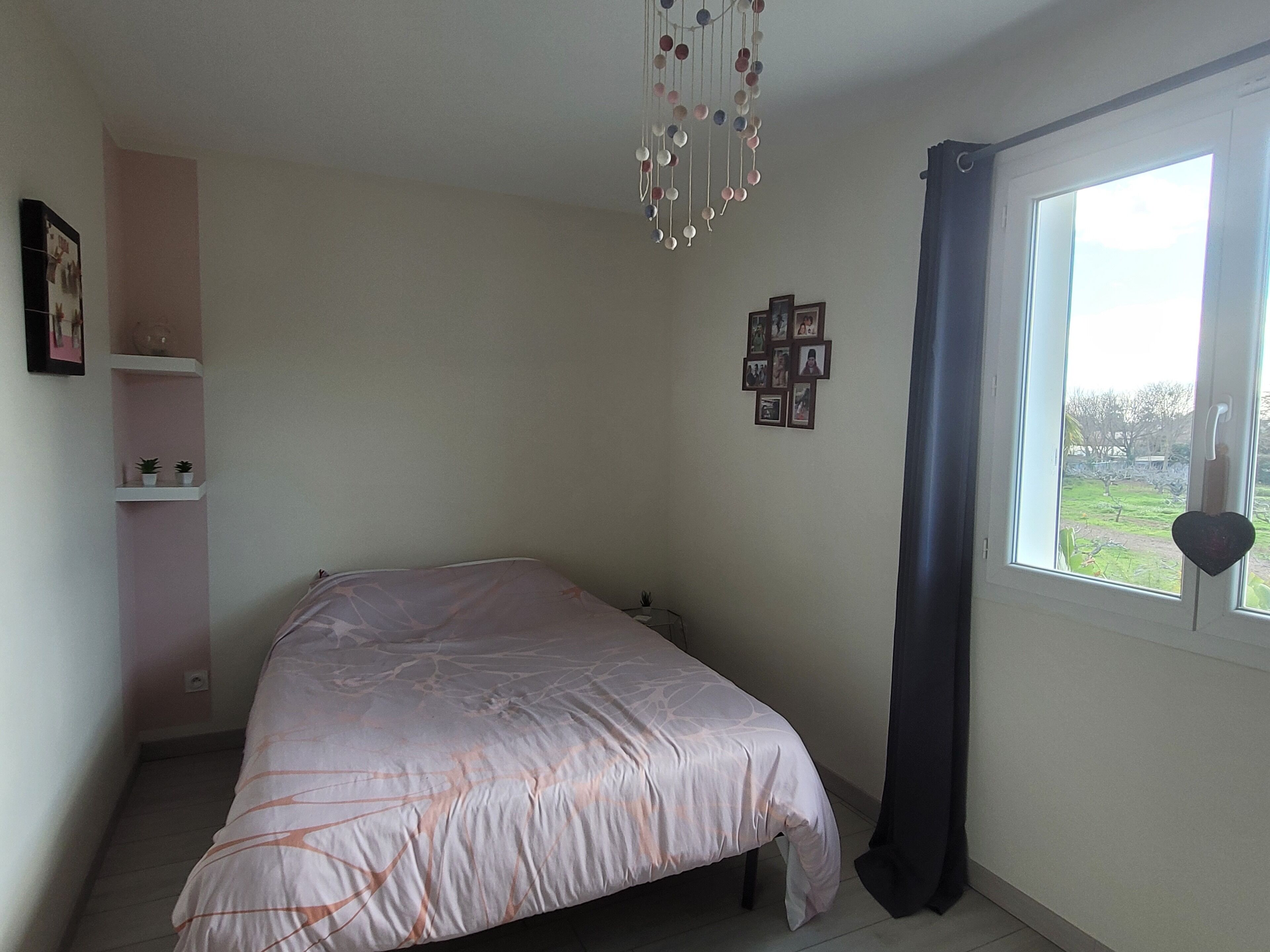 A bedroom with a bed, a window with curtains, a wall-mounted shelf with plants, and a wall with framed pictures.