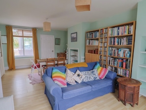 A living room with a blue sofa, a wooden bookshelf, a dining table with chairs, and a window with curtains. - 20 Lym Close (Lyme Regis)