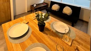 Uma área de jantar com mesa de madeira posta para quatro pessoas, um vaso de planta e um sofá preto com almofadas.