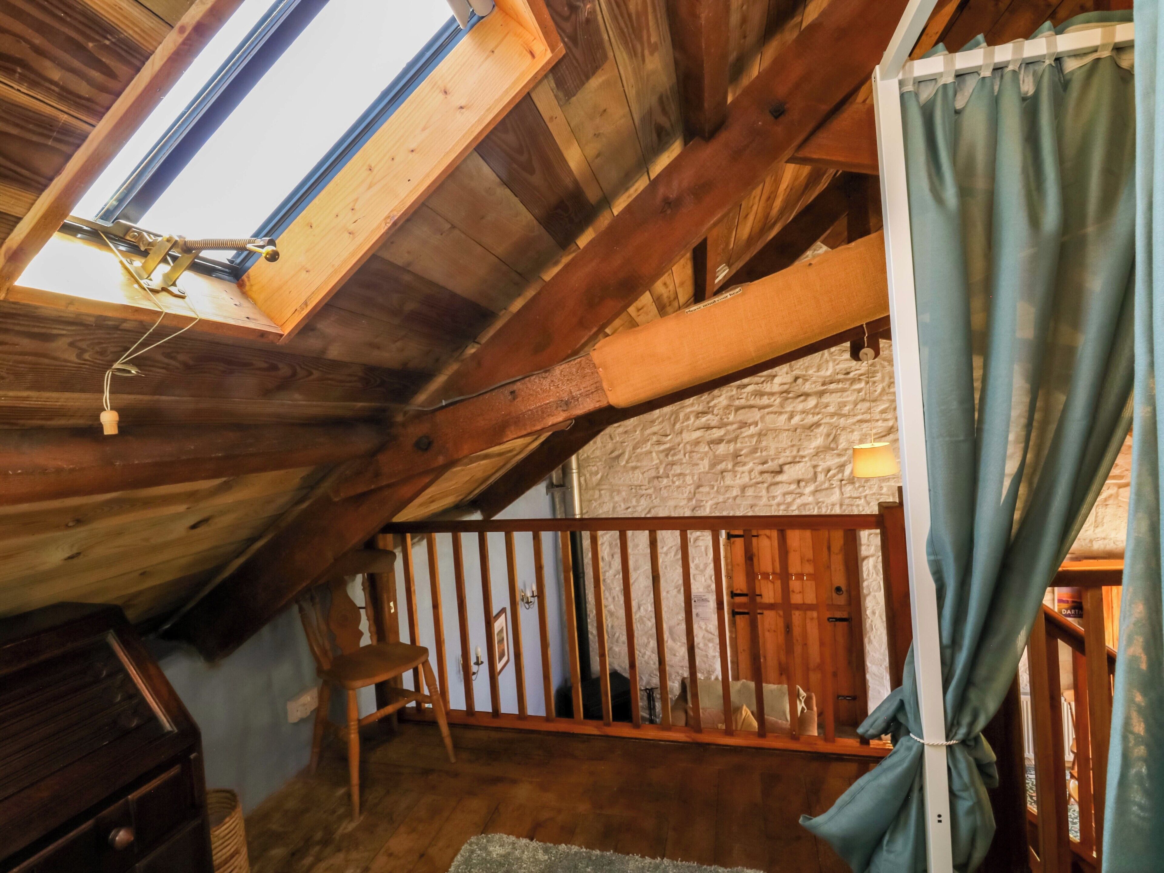 A rustic attic room with wooden beams, a sloped ceiling, a wooden chair, a small table, and a view through a window.