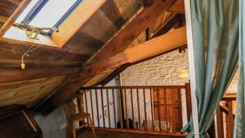 A rustic attic room with wooden beams, a sloped ceiling, a wooden chair, a small table, and a view through a window.