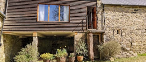 Une maison en pierre à deux étages, dotée d’une façade en bois, d’un balcon et de plantes en pot dans le jardin.