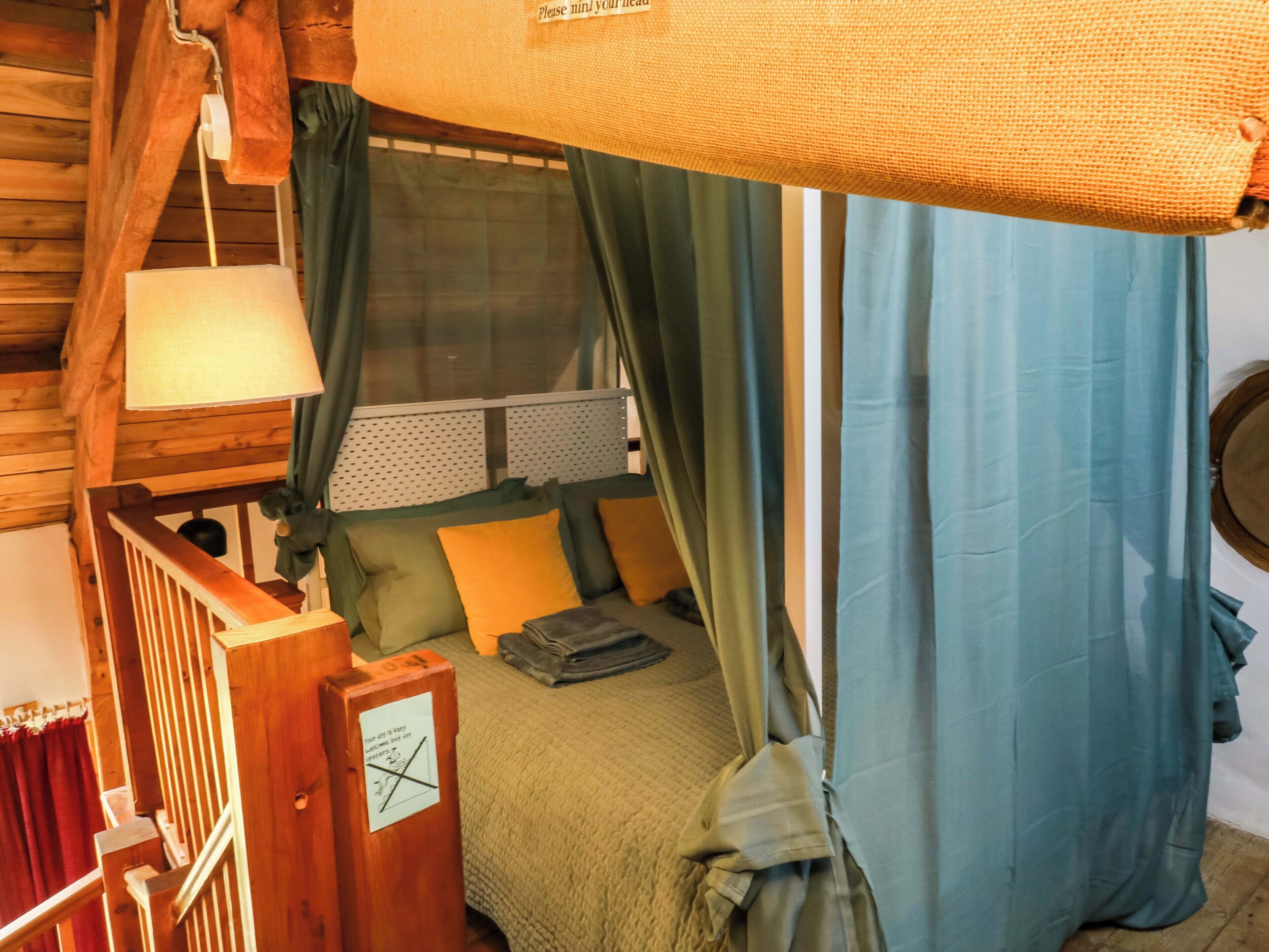 Una habitació acollidora amb un llit, una finestra amb cortines, una escala de fusta i un rellotge de paret.