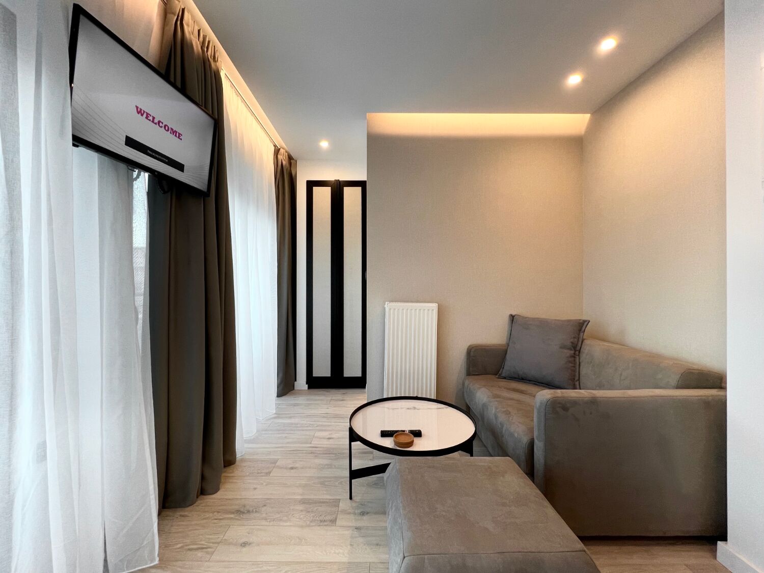 A modern living room with a sofa, ottoman, and a small table with a remote control. A TV is mounted on the wall, and there is a radiator in the corner.