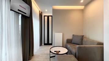 A modern living room with a sofa, ottoman, and a small table with a remote control. A TV is mounted on the wall, and there is a radiator in the corner.