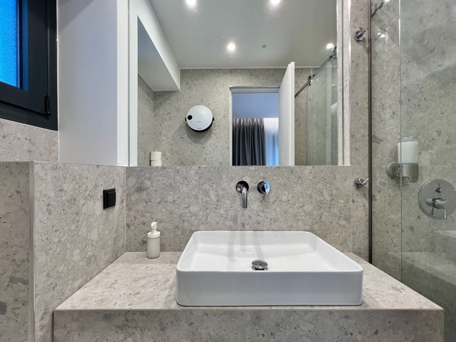 A modern bathroom with a rectangular sink, two faucets, a soap dispenser, and a handheld mirror.