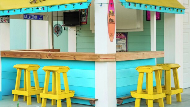 A colorful beach-themed kiosk with a surfboard, yellow stools, and a sign reading "GALVESTON AVE 4000". - Your Coastal Getaway:3BR/2BA Beach Retreat (Bolivar Peninsula)