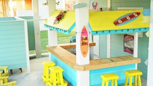 A beach-themed bar area with a surfboard decoration, yellow stools, and a sign reading "HEFTEN-A". - Your Coastal Getaway:3BR/2BA Beach Retreat (Bolivar Peninsula)