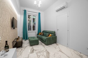 Un salon avec un canapé vert, des rideaux turquoise, un téléviseur à écran plat, une petite table avec une bouteille et des verres, et une porte blanche.