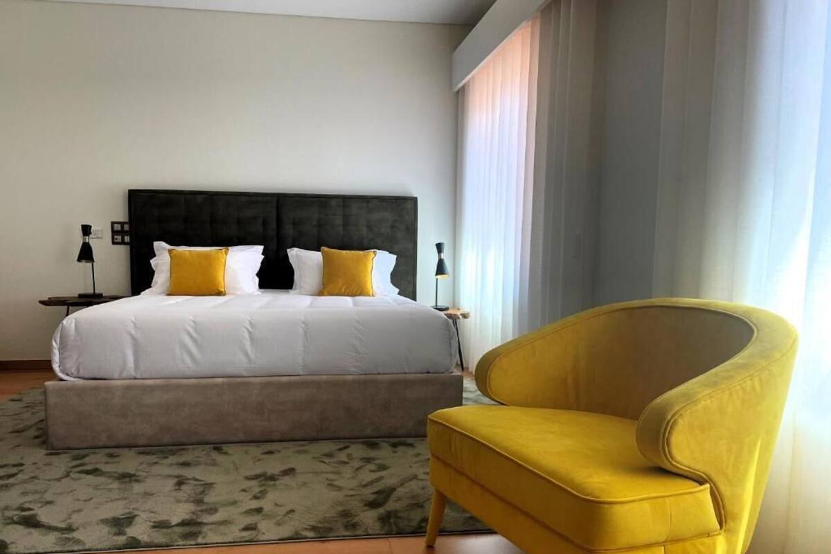 Una habitación de hotel con una cama, una mesita de noche, una lámpara, un sillón amarillo y una ventana con cortinas.