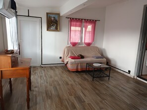 Ein Zimmer mit Holzboden, einer Couch mit Decke, einem kleinen Tisch, einem Bild an der Wand und einem Fenster mit Vorhängen.
