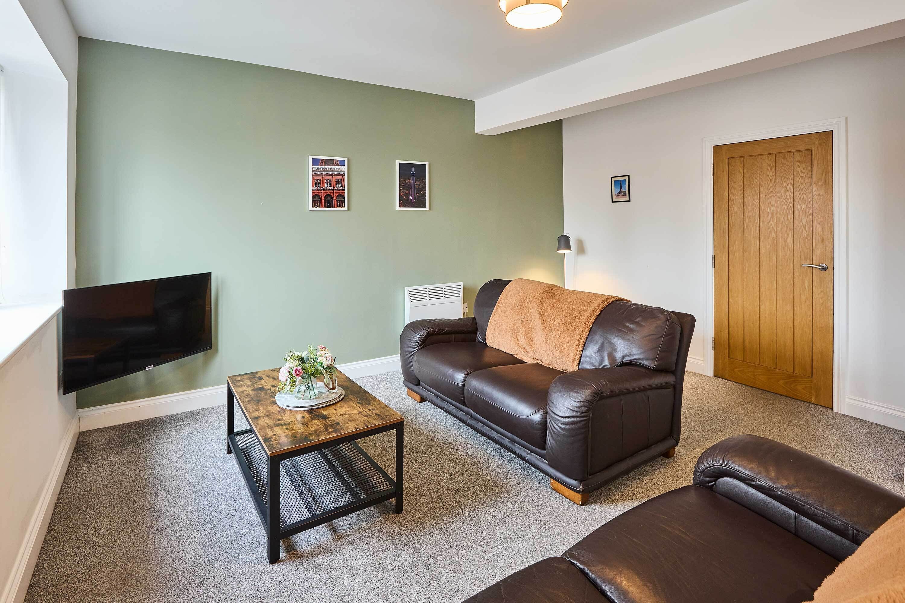 A living room with a brown leather sofa, a coffee table, and a television. There are two framed pictures on the wall and a door in the background.