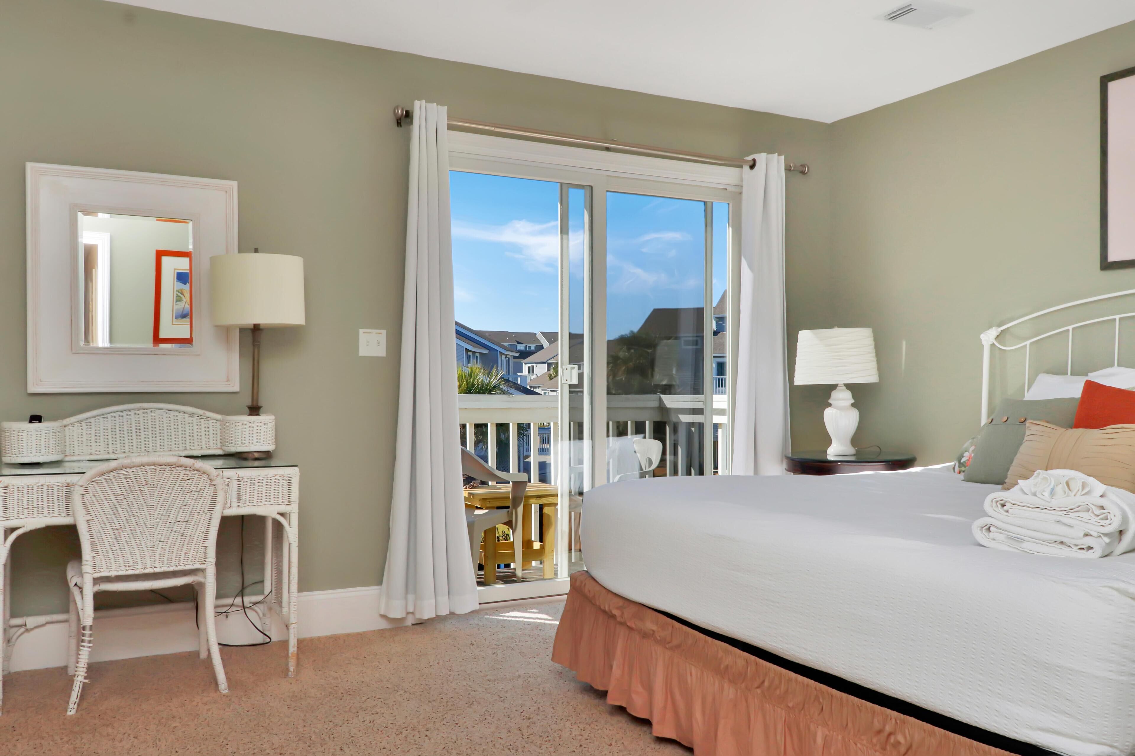 A hotel room with a bed, a desk, a chair, and a balcony with a view of houses.