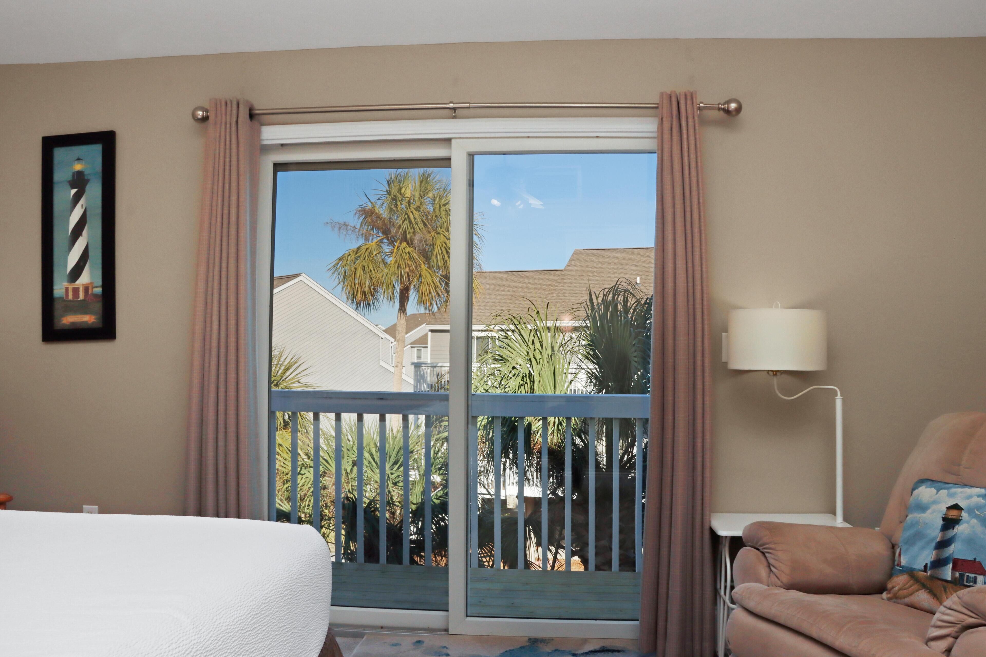 A hotel room with a bed, a chair, a lamp, and a view of palm trees and a building through a sliding glass door.