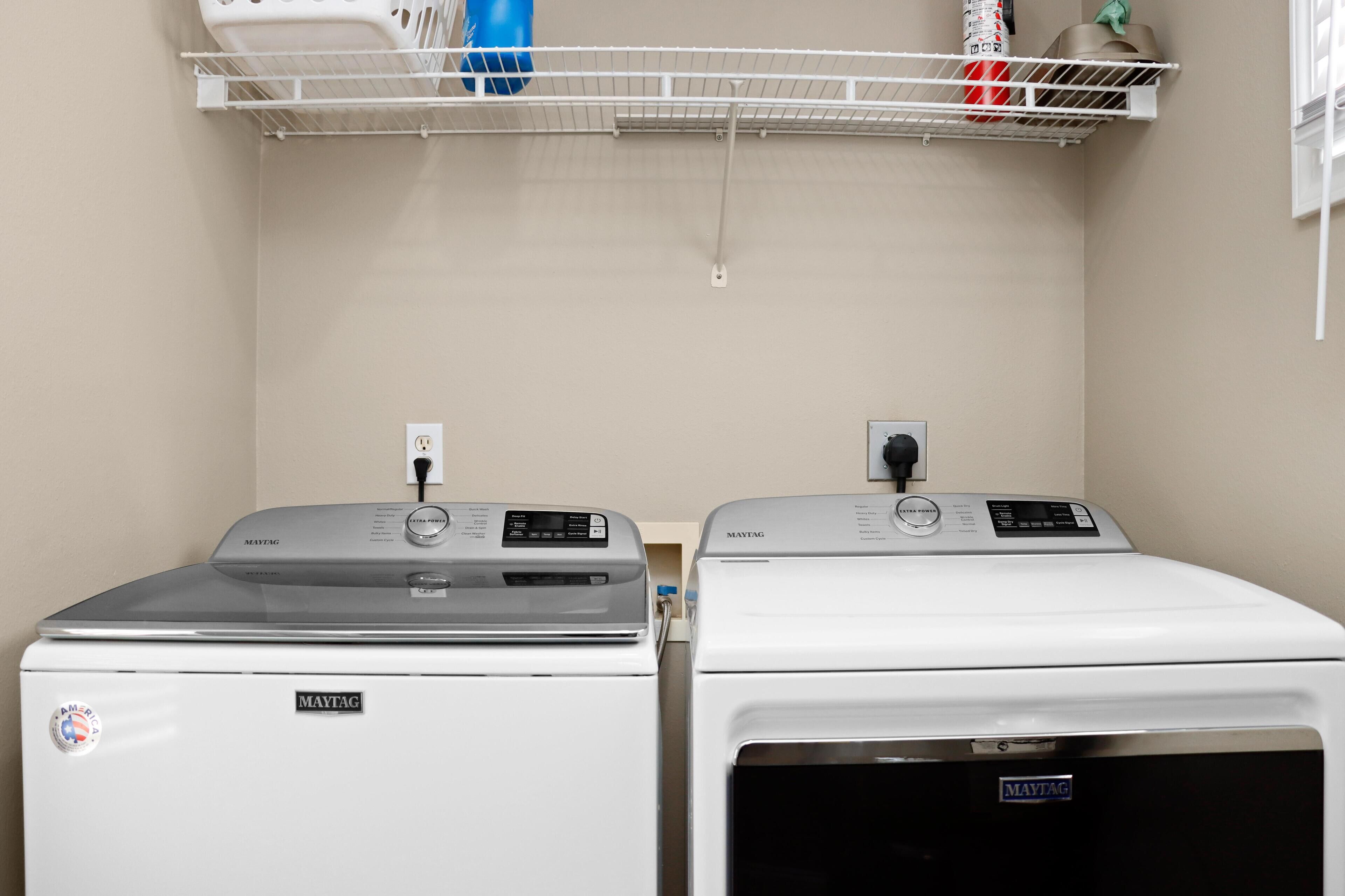 Two Maytag washing machines in a laundry room with a white shelf above them.