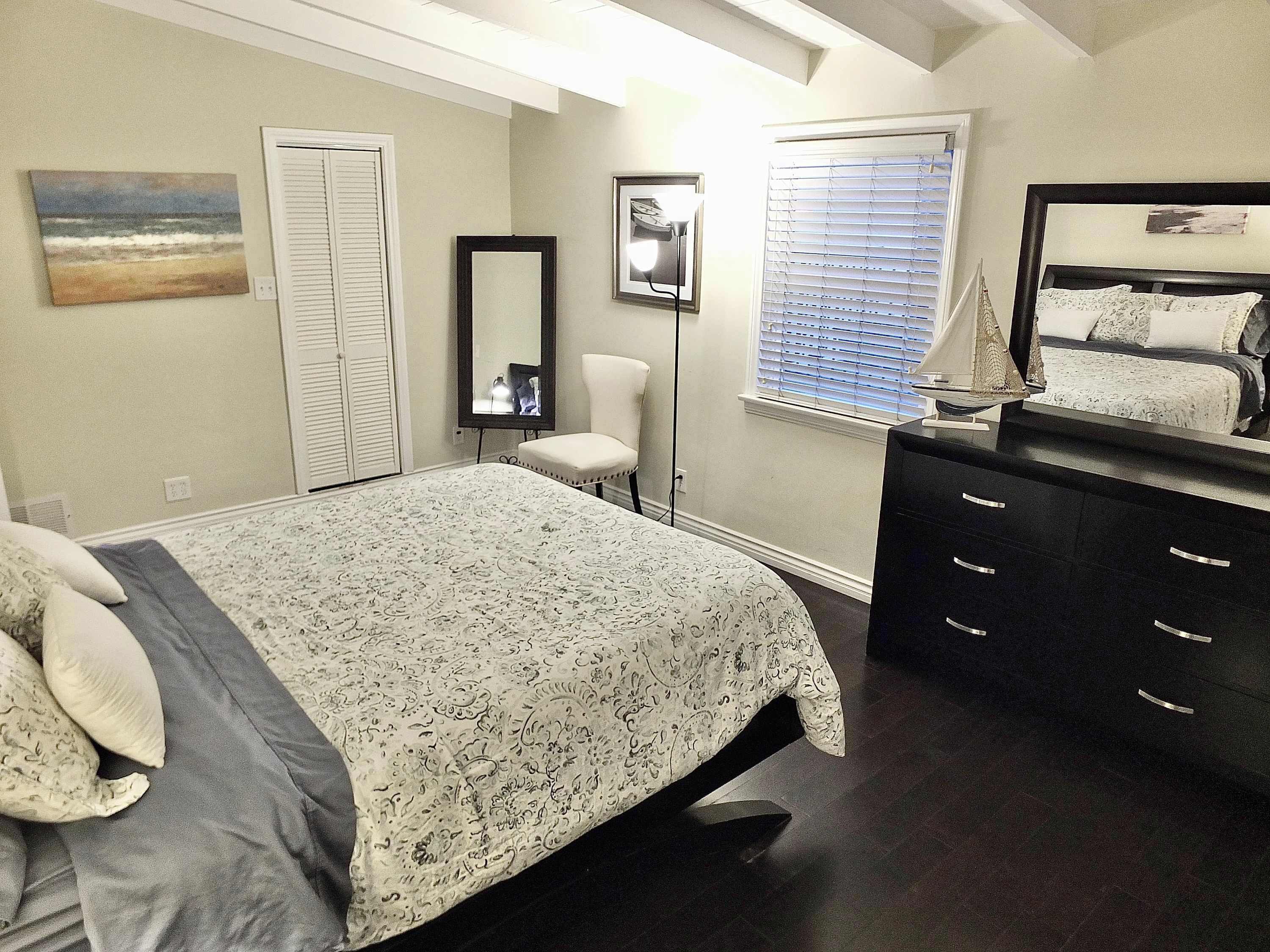 Un dormitorio con cama, cómoda, silla, ventana con persianas, espejo y un cuadro en la pared.