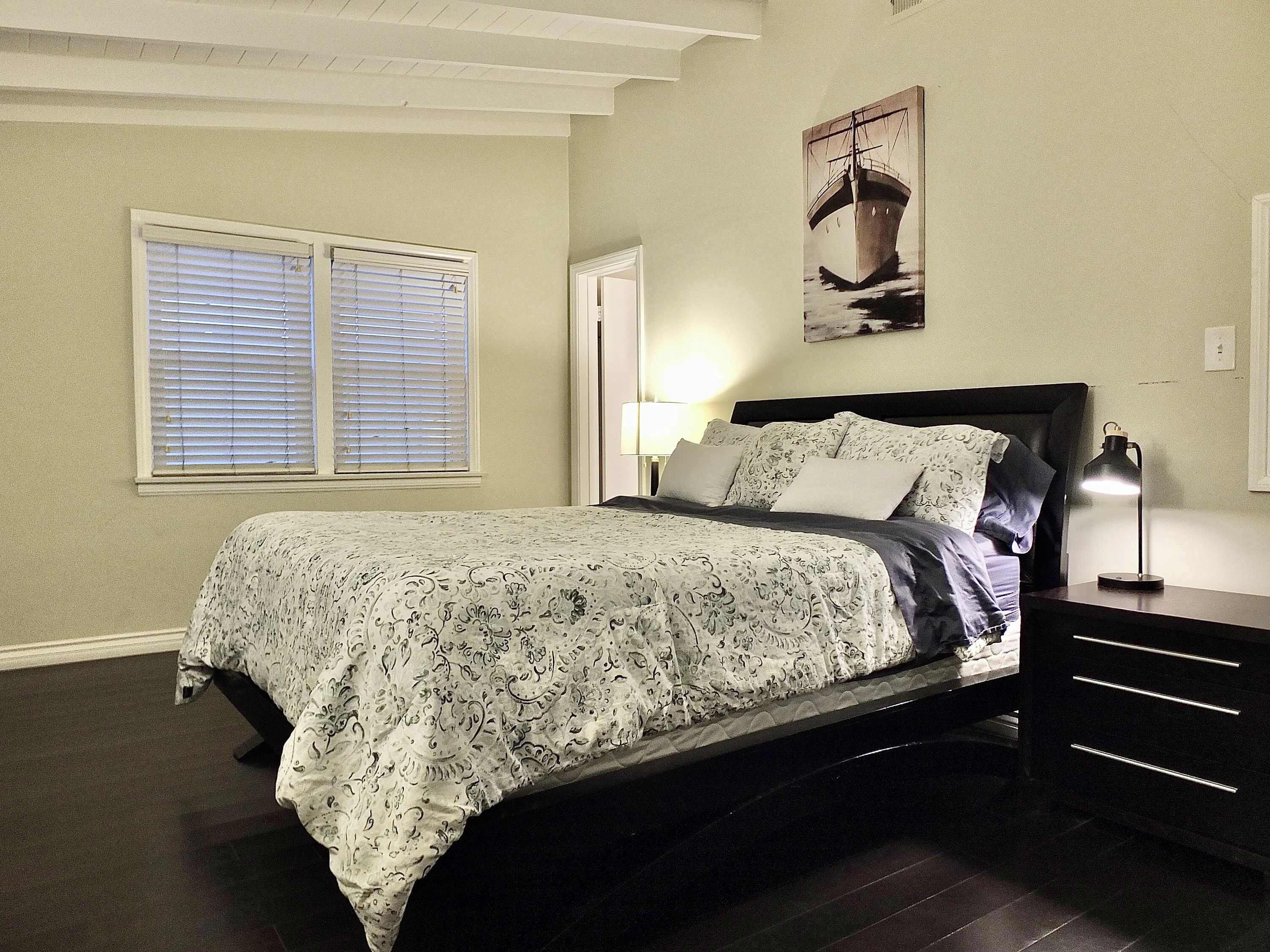 A bedroom with a bed, a nightstand, a lamp, a window with blinds, and a wall-mounted picture.
