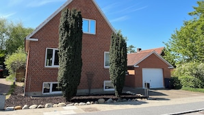 A two-story brick house with a gabled roof, two tall cypress trees in front, a garage, and a well-maintained lawn. - Struer Family House | 600m to Beach, Garden with Trampoline & Play Area (Struer)