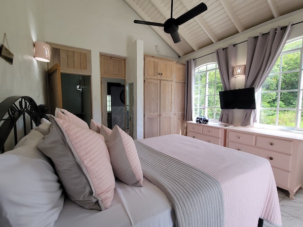 A bedroom with a bed, a ceiling fan, a TV, a window with curtains, and wooden cabinets.