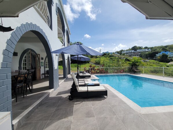 Una piscina con sillas de descanso y sombrillas, un patio cubierto con asientos, y una vista de un paisaje verde exuberante al fondo.