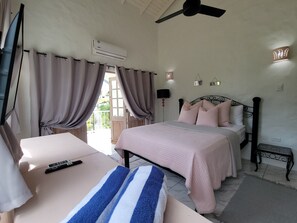 Un dormitorio con cama, mesita de noche, cortinas, unidad de aire acondicionado, ventilador de techo y una pequeña mesa.
