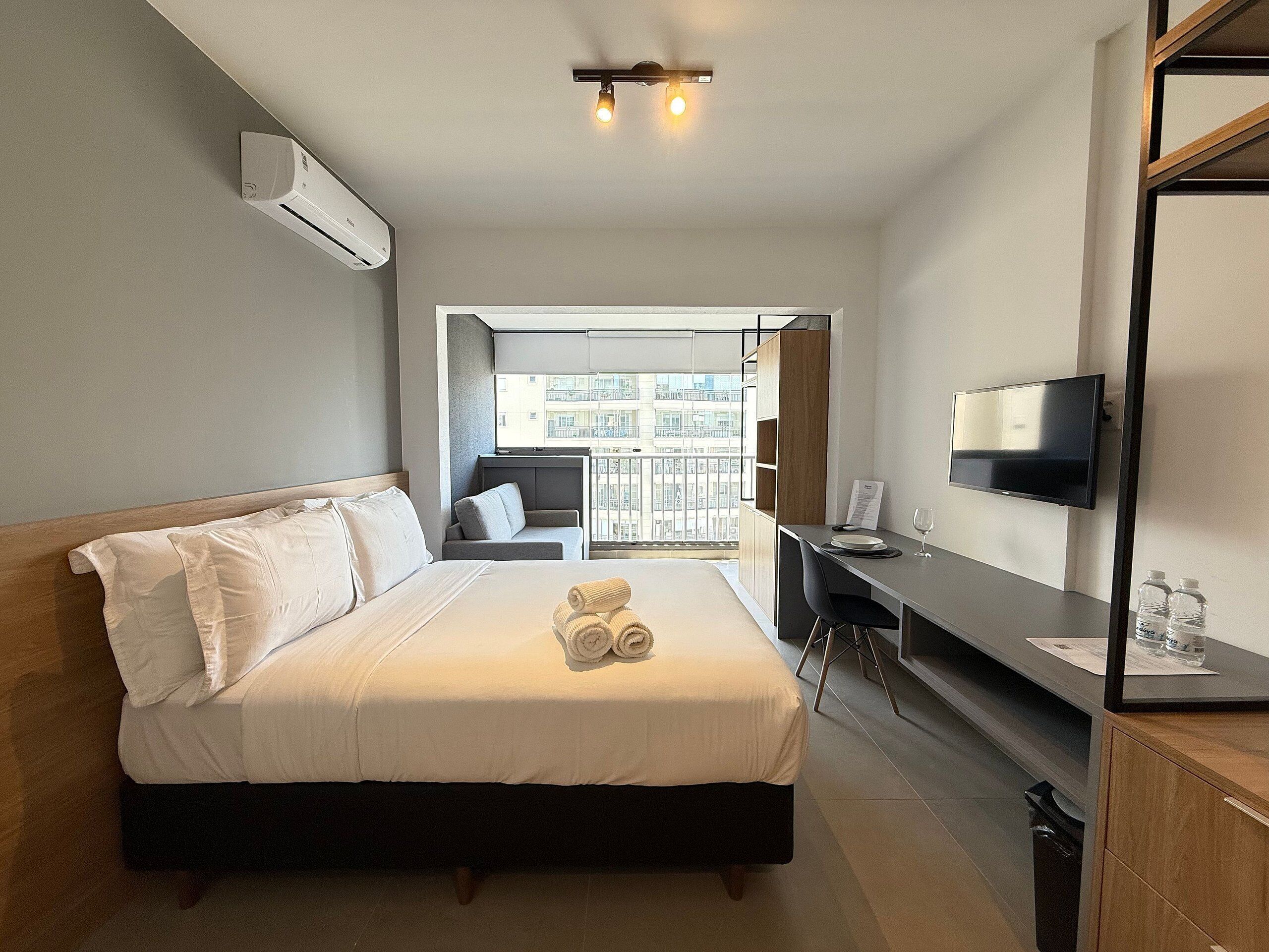 A hotel room with a bed, a desk, a TV, and a balcony with a city view.