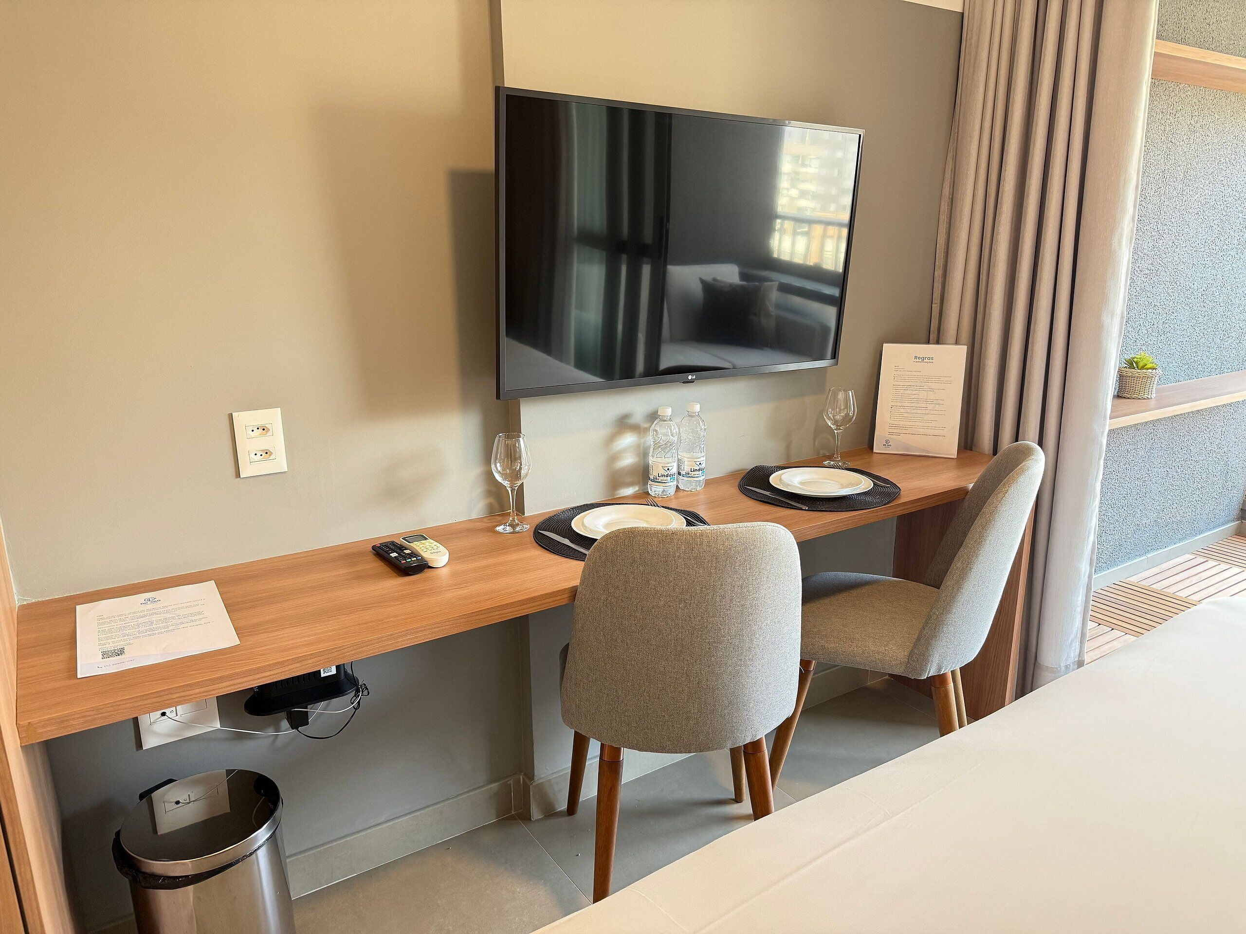 A hotel room with a wooden desk, two chairs, a television, a trash bin, and a window with curtains.