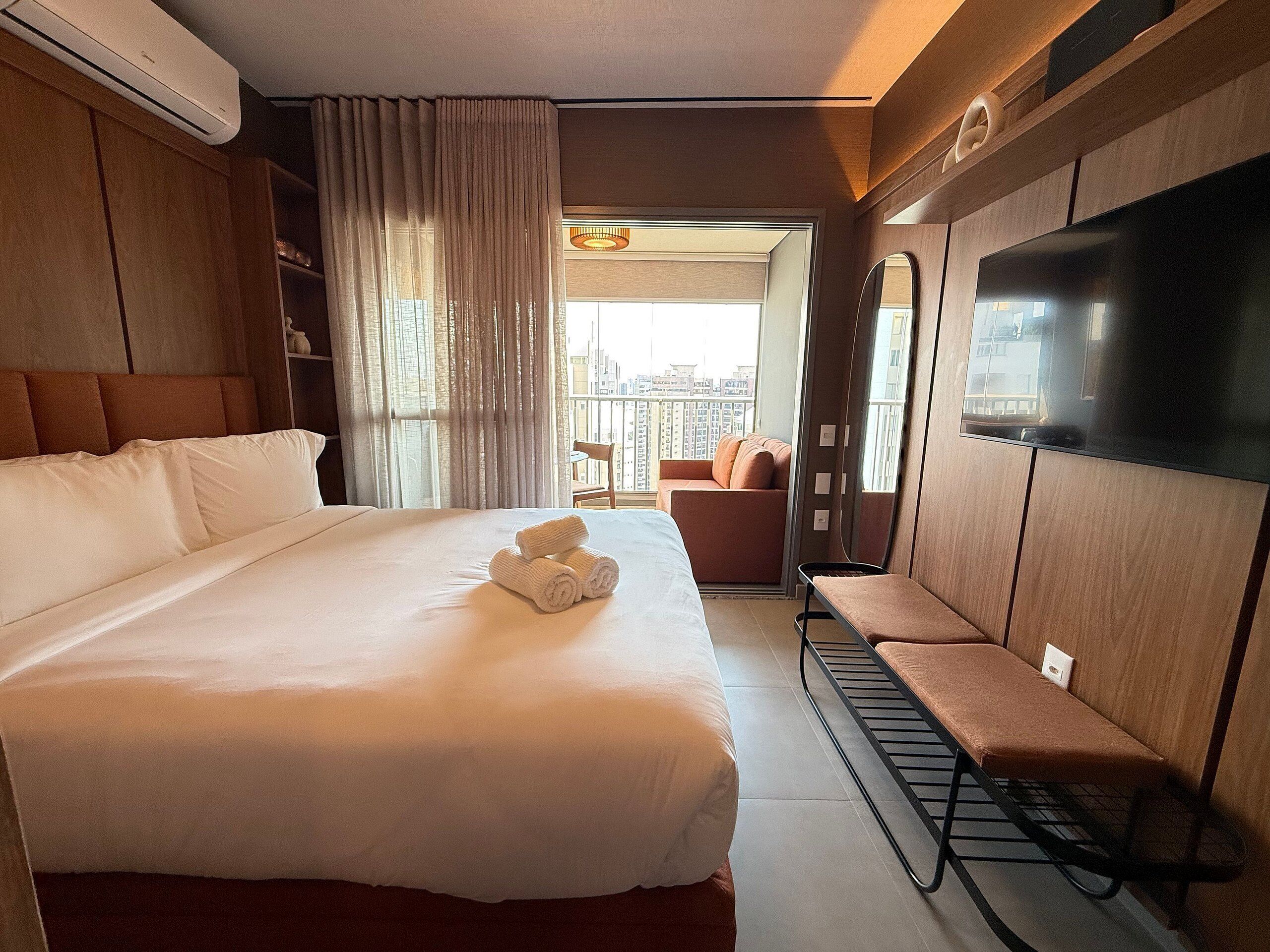 A hotel room with a large bed, a TV, a seating area, and a view of the city.