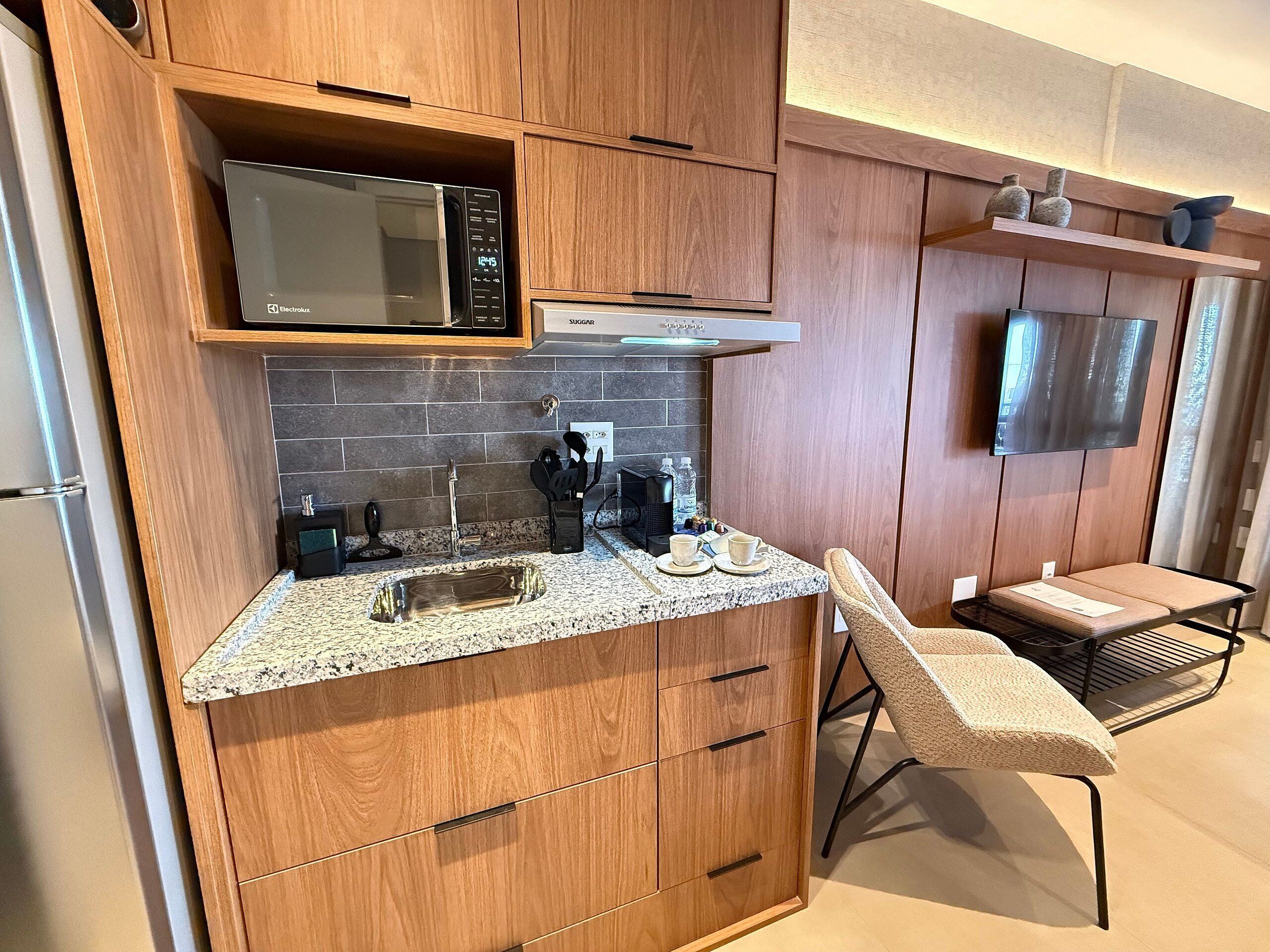 Cozinha compacta com micro-ondas, pia e cafeteira. Possui um pequeno espaço de estar com uma cadeira e uma mesa.