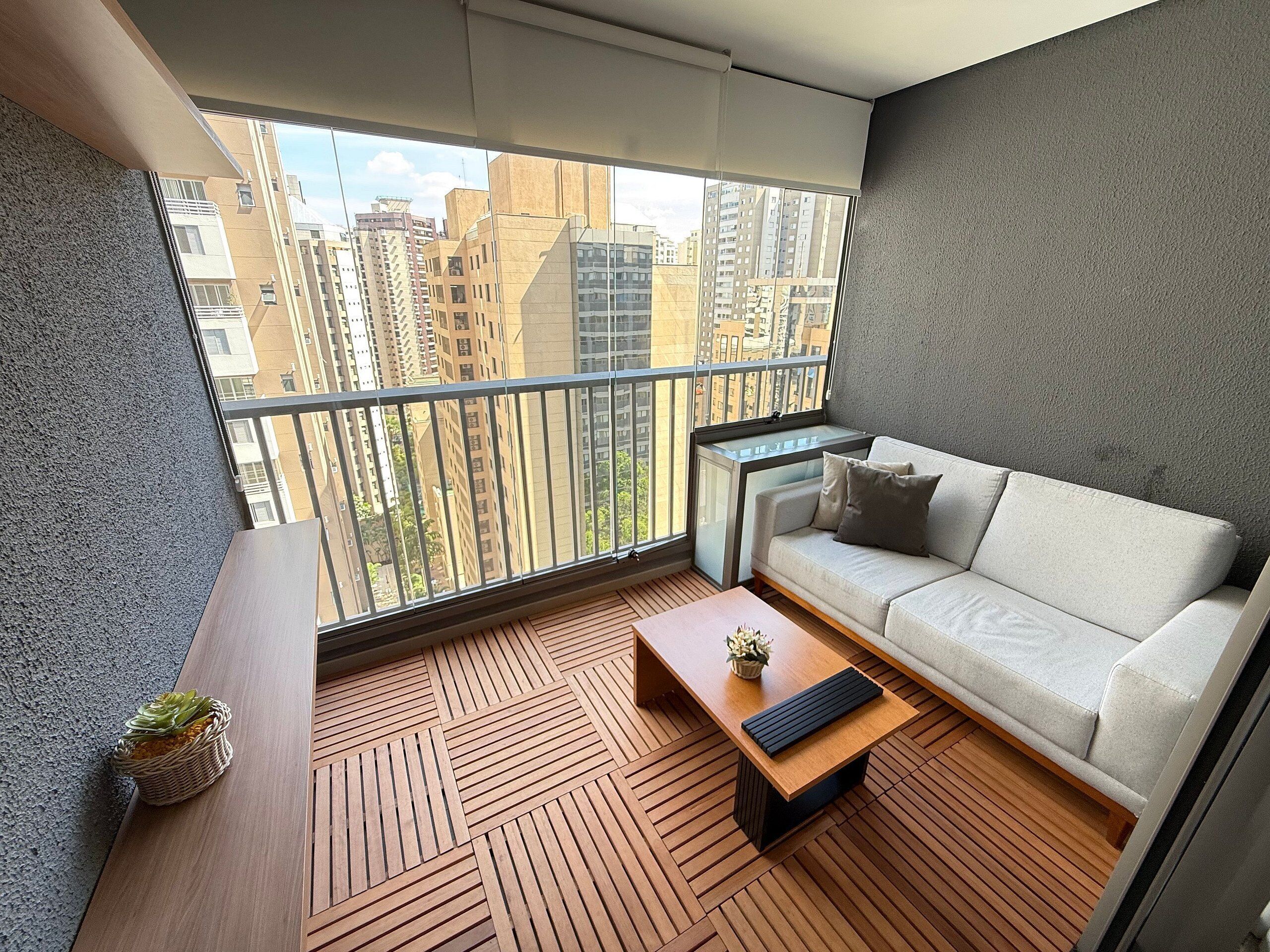 Uma varanda moderna com sofá e mesa de centro, com vista para a cidade através de amplas janelas.