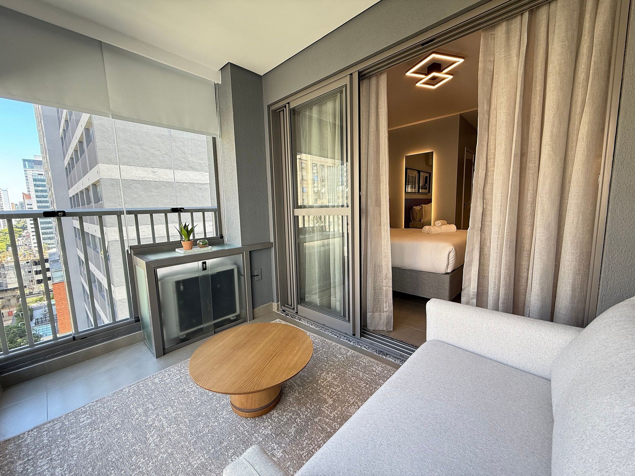 A modern hotel room with a balcony, a round wooden coffee table, a sofa, and a bed with curtains.