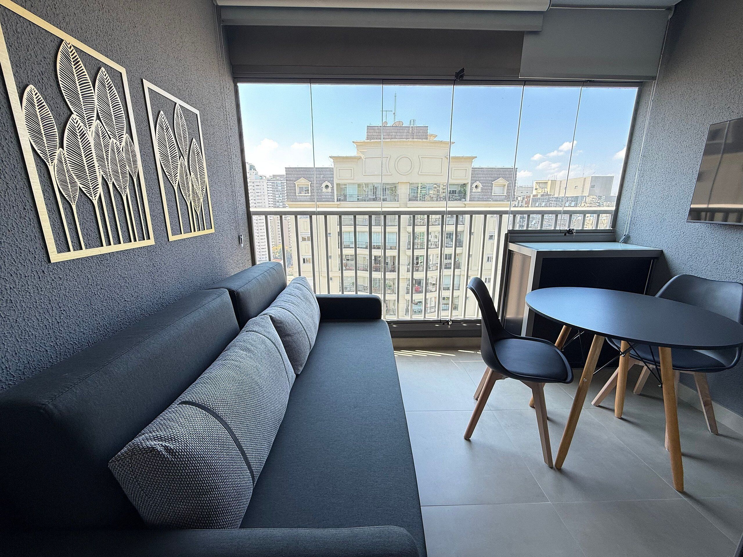 A modern living room with a grey sofa, a small round table, and chairs, a balcony with a view of buildings.