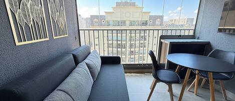 A modern living room with a grey sofa, a small round table, and chairs, a balcony with a view of buildings.