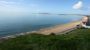 Egy tengerparti kilátás, ahol homokos strand, nyugodt tenger és egy fűvel borított domb látható.